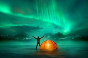 Man standing with out-stretched arms beside an orange tent looking towards northern lights over the mountains