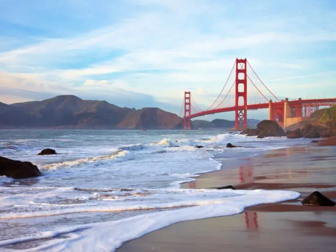 Red bridge crossing a body of water with waves crashing on a sandy beach in the foreground