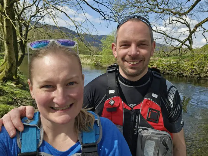 A man and a woman in canoe gear and buoyancy aids with a river and trees in the background