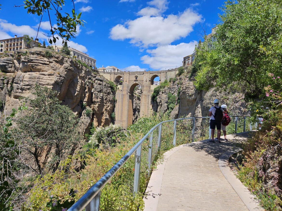 Paved walkway with 2 people wearing helmets, and a stone bridge across a gorge in the background