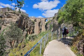 Paved walkway with 2 people wearing helmets, and a stone bridge across a gorge in the background