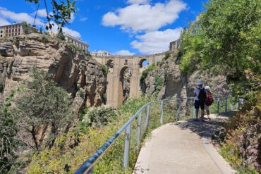 Paved walkway with 2 people wearing helmets, and a stone bridge across a gorge in the background