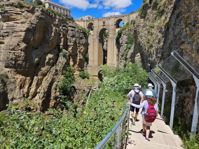 Big stone bridge across a deep gorge, with a path leading to it and 3 people wearing white helmets